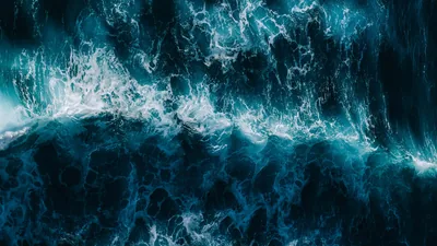 An overhead view of a wave folding over itself, foam veins spreading across petrol-blue water with the darkest patches pooled in both lower corners.