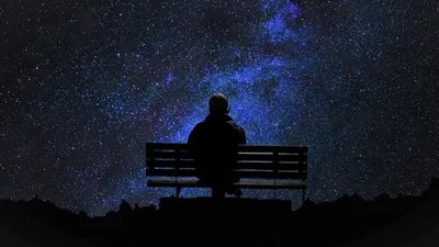 Seen from behind, someone sits on a wooden bench beneath a violet band of stars, the ground below the bench fading to full black.