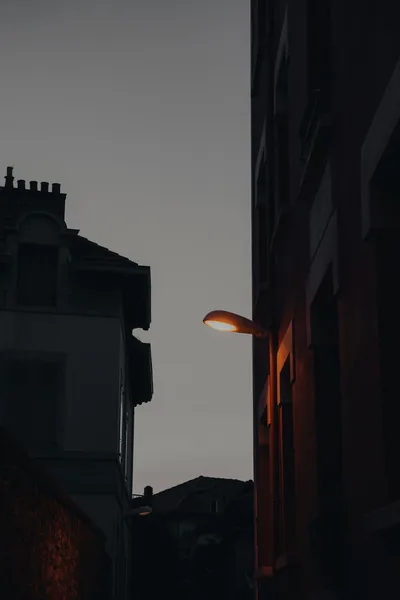 An orange wall lamp juts from a red-brick building at dusk, a strip of flat grey sky running down the middle of this tall portrait frame.
