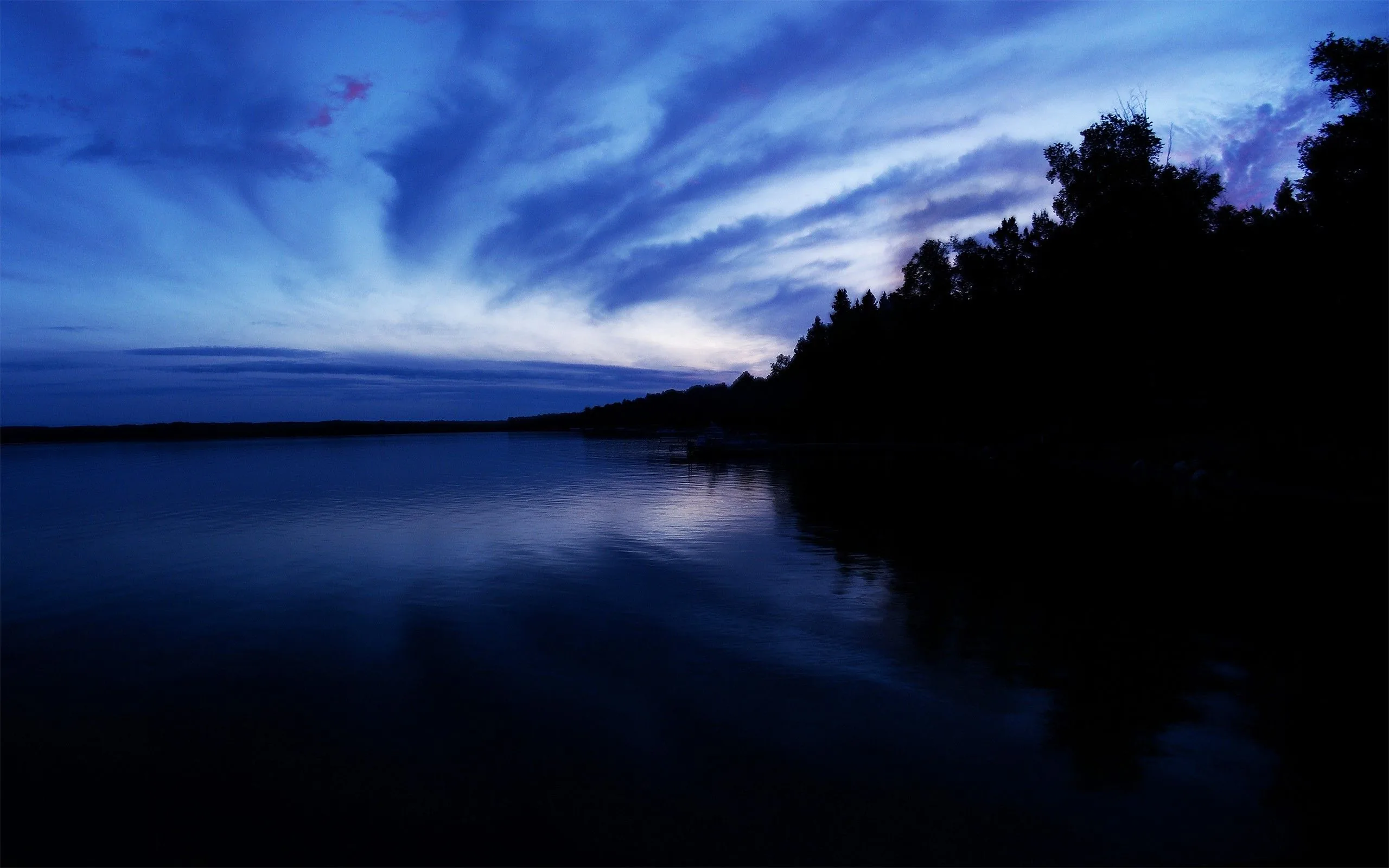 Streaked cloud in deep blue hangs over still water, a black treeline running down the right bank and the far shore flattened to a single line.