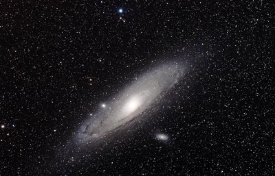 A spiral galaxy seen at an angle, its pale core burning through dusty arms while a smaller companion sits just below it in the star field.