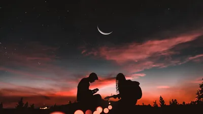 Two people sit close on a ridge under a thin crescent moon, the horizon burning orange below layered cloud and a scatter of stars above.
