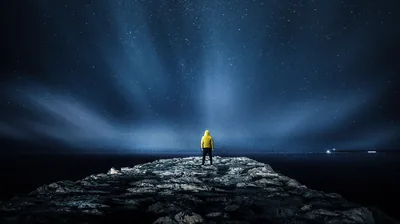 Blue beams fan upward from the sea horizon behind a man in a yellow jacket on the rocks, faint ship lights dotting the far right.