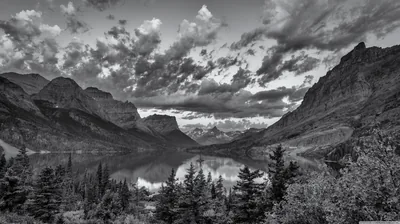 A still lake runs between steep cliffs with pines in the foreground and cloud stacked over the far peaks.