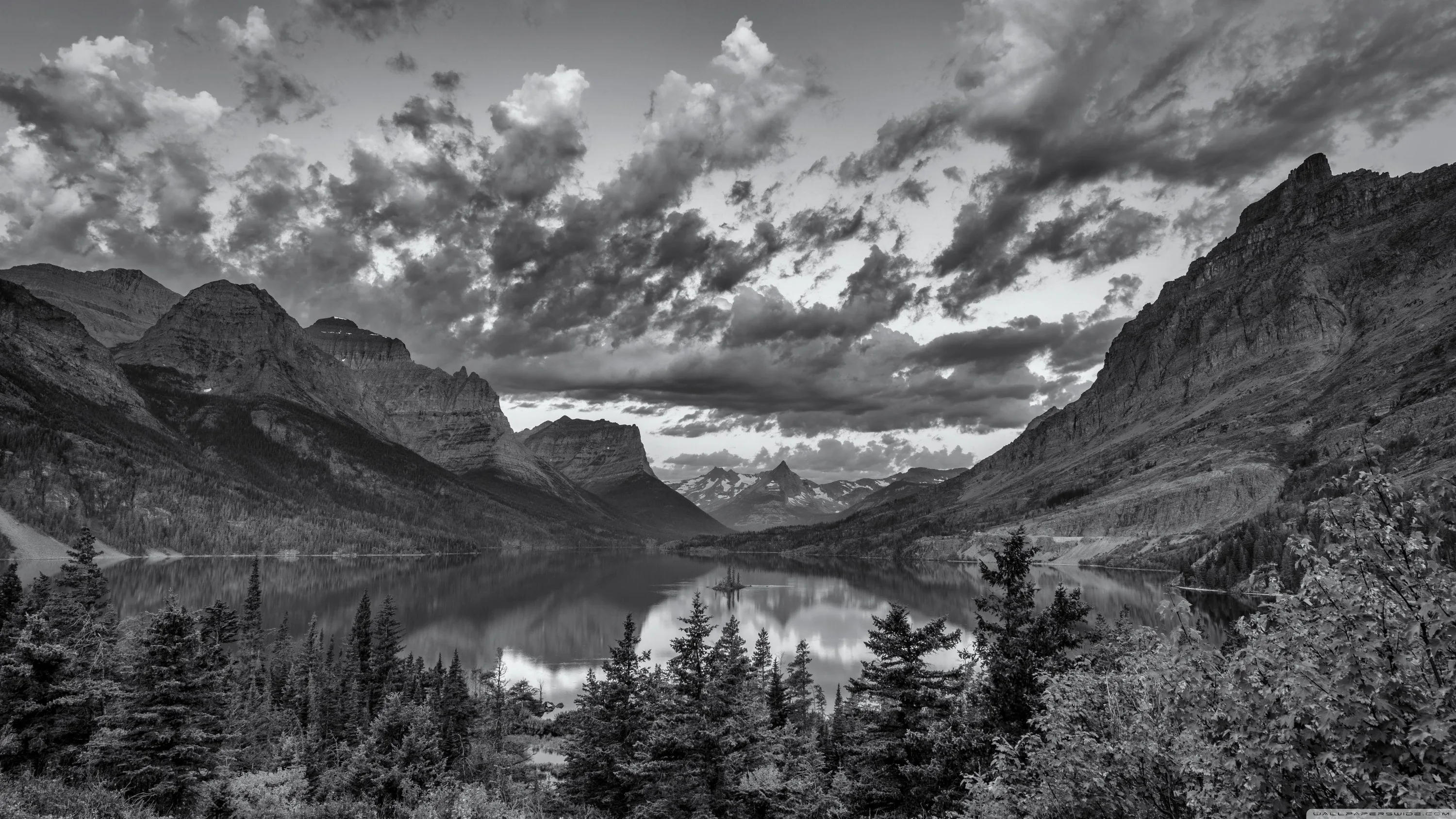 A still lake runs between steep cliffs with pines in the foreground and cloud stacked over the far peaks.