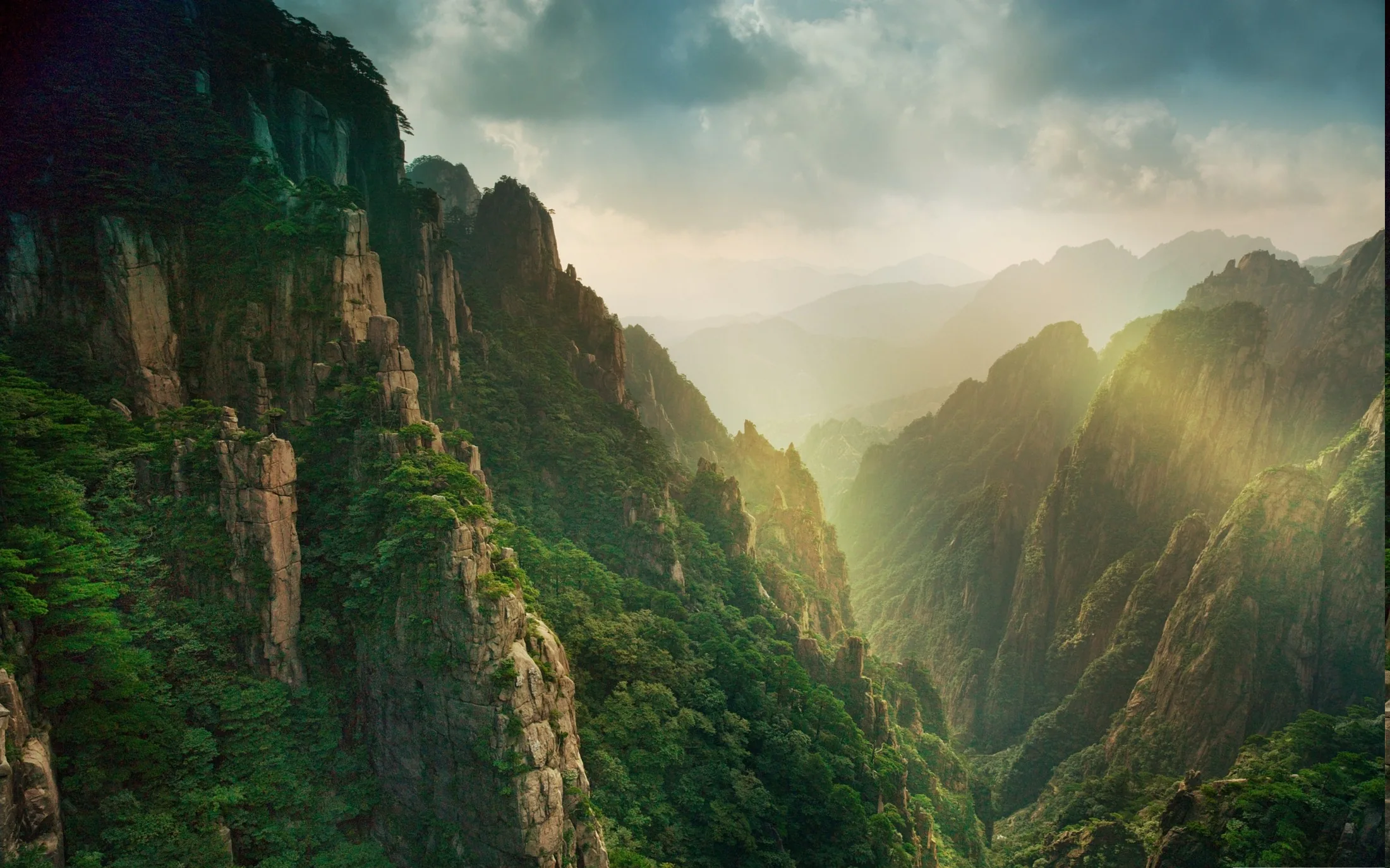 Rays fan through haze between forested stone pinnacles in a Chinese canyon, the bright break in the clouds drawing the eye to the centre of the frame.