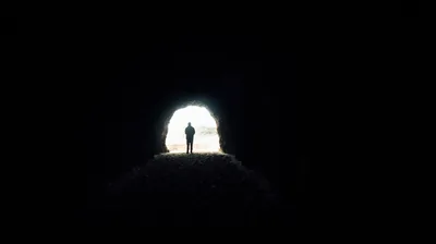 Everything is black except the ragged mouth of a tunnel, where a figure stands small against white daylight just right of centre - flat black suits OLED.