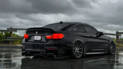 Three-quarter view from behind, the M4's carbon diffuser and quad pipes over a wet surface with grey cloud and a motorway barrier in the distance.