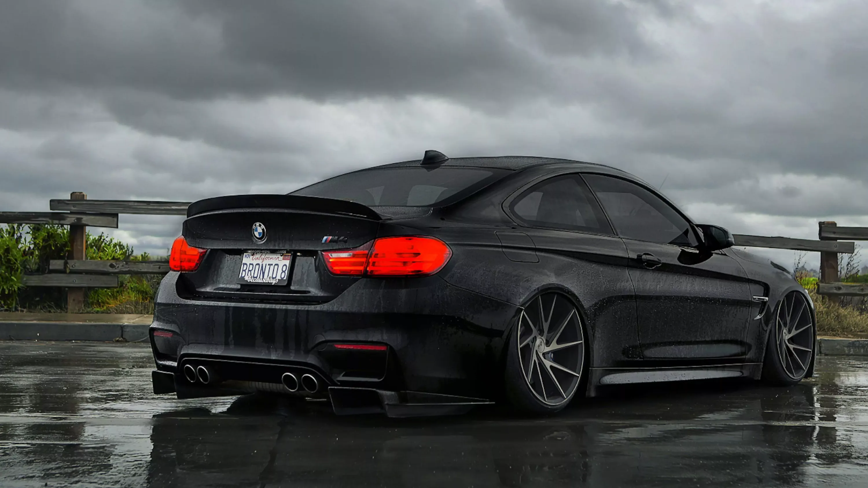 Three-quarter view from behind, the M4's carbon diffuser and quad pipes over a wet surface with grey cloud and a motorway barrier in the distance.