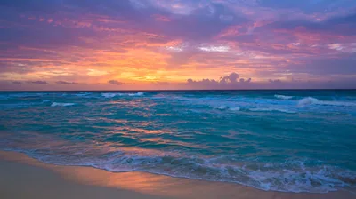 Pink and lavender clouds hang over rolling turquoise waves, the wet beach in front reflecting the sky's colour in a soft sheen.