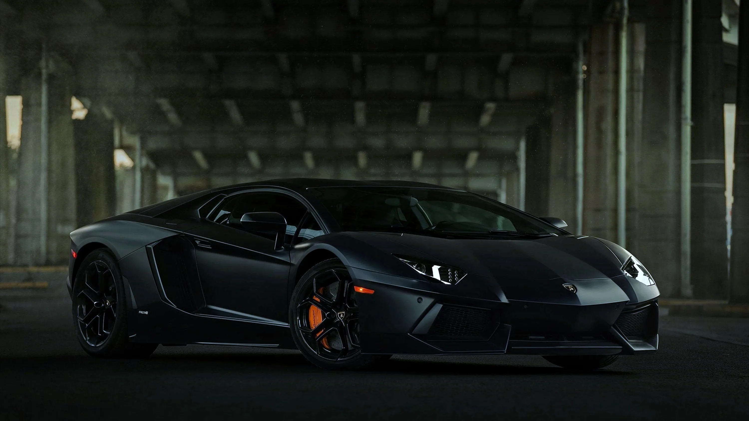 Matte dark grey Aventador parked in the shade of a bridge, orange brake calipers glowing behind black wheels and the pillars fading out behind it.