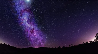 The galactic core burns pink and purple above a black tree line, with warm light from a distant town spilling in from both sides.