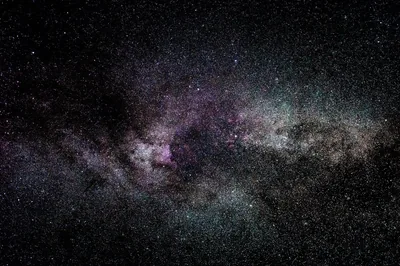 A wide astrophotograph of the galactic plane, dark dust webs cutting through pale star fields with a magenta emission region left of centre.