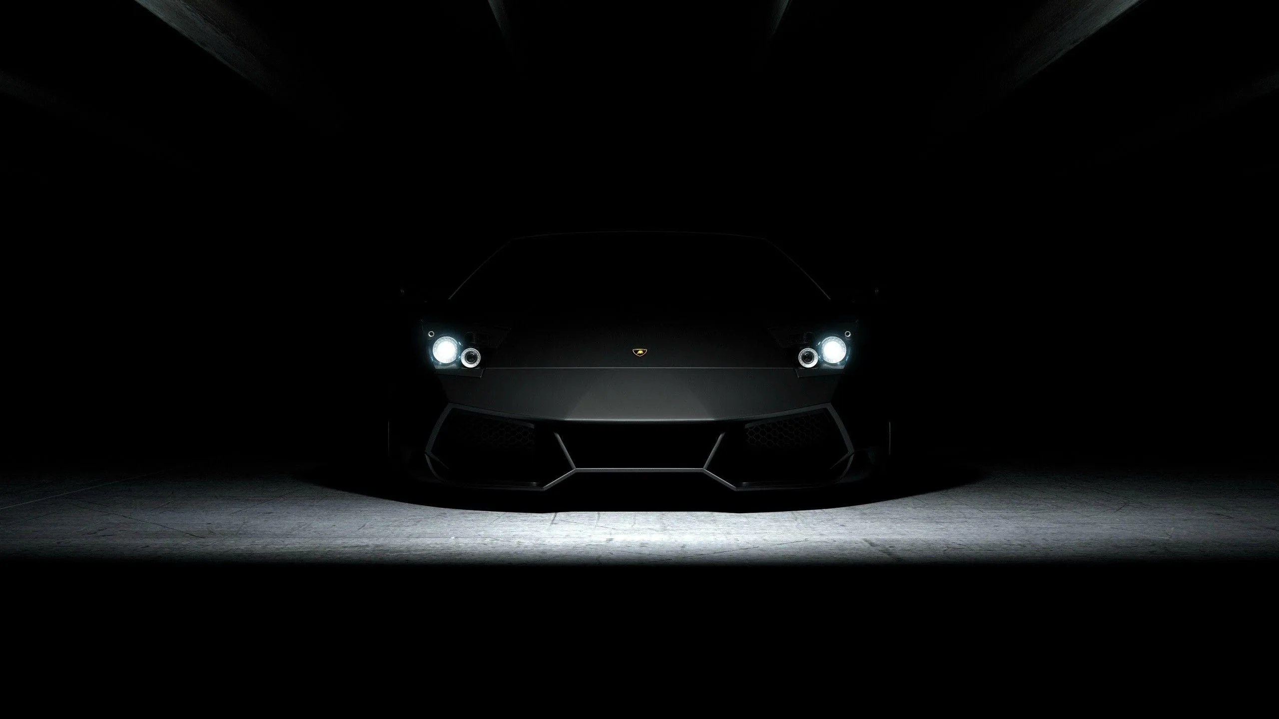 A low sports car sits nose-on in a black space, only its headlights and the light pooling under it visible.
