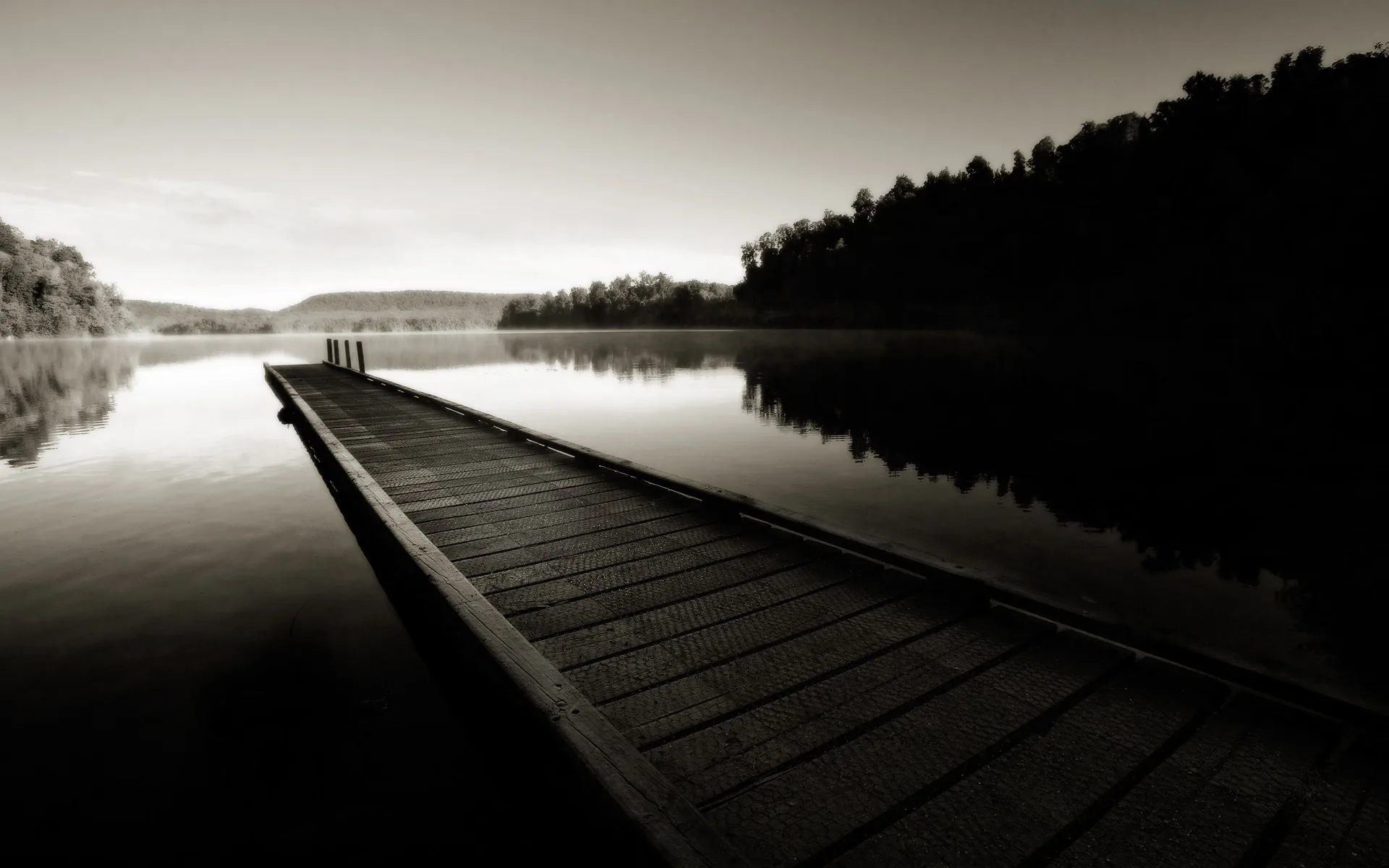 A weathered jetty runs out into flat water at dawn, the treeline and hills rendered in warm sepia.