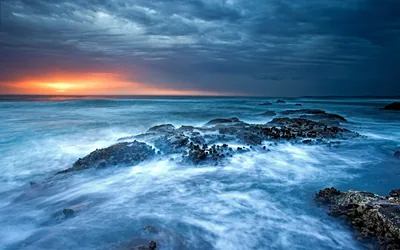 Blurred water flows like fog over a dark reef beneath a heavy grey sky, one strip of orange sunset burning at the far left horizon.