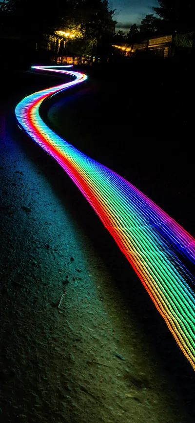 A long-exposure rainbow ribbon of light snakes up a dark garden path at night, the grass around it staying black enough for app icons.