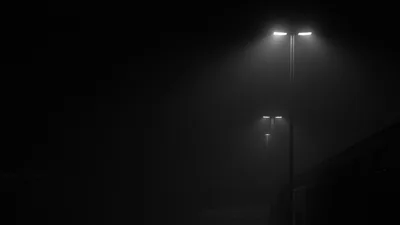 Two lamp posts glow through heavy fog on an empty street, a low wall just visible at the right edge.