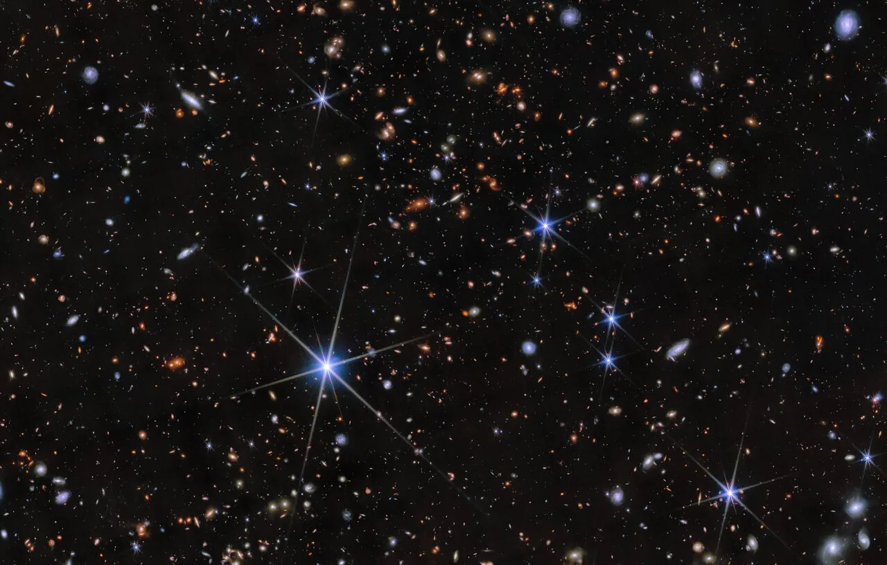 Small orange and white galaxies scattered across deep black, one foreground star throwing six diffraction spikes near the lower left.