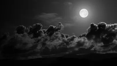 A bright moon clears a bank of heavy cloud in monochrome, the land below reduced to a flat silhouette.