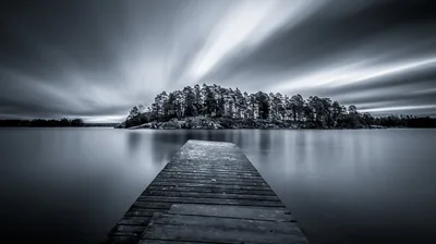A long exposure pulls the clouds into streaks above a small wooded island, the weathered pier running straight out from the bottom edge.