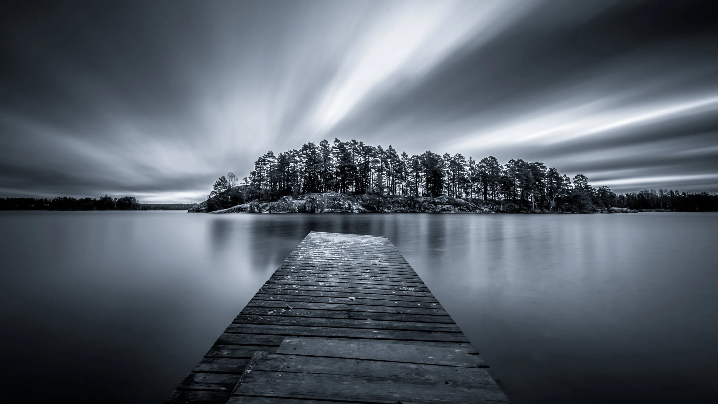 A long exposure pulls the clouds into streaks above a small wooded island, the weathered pier running straight out from the bottom edge.