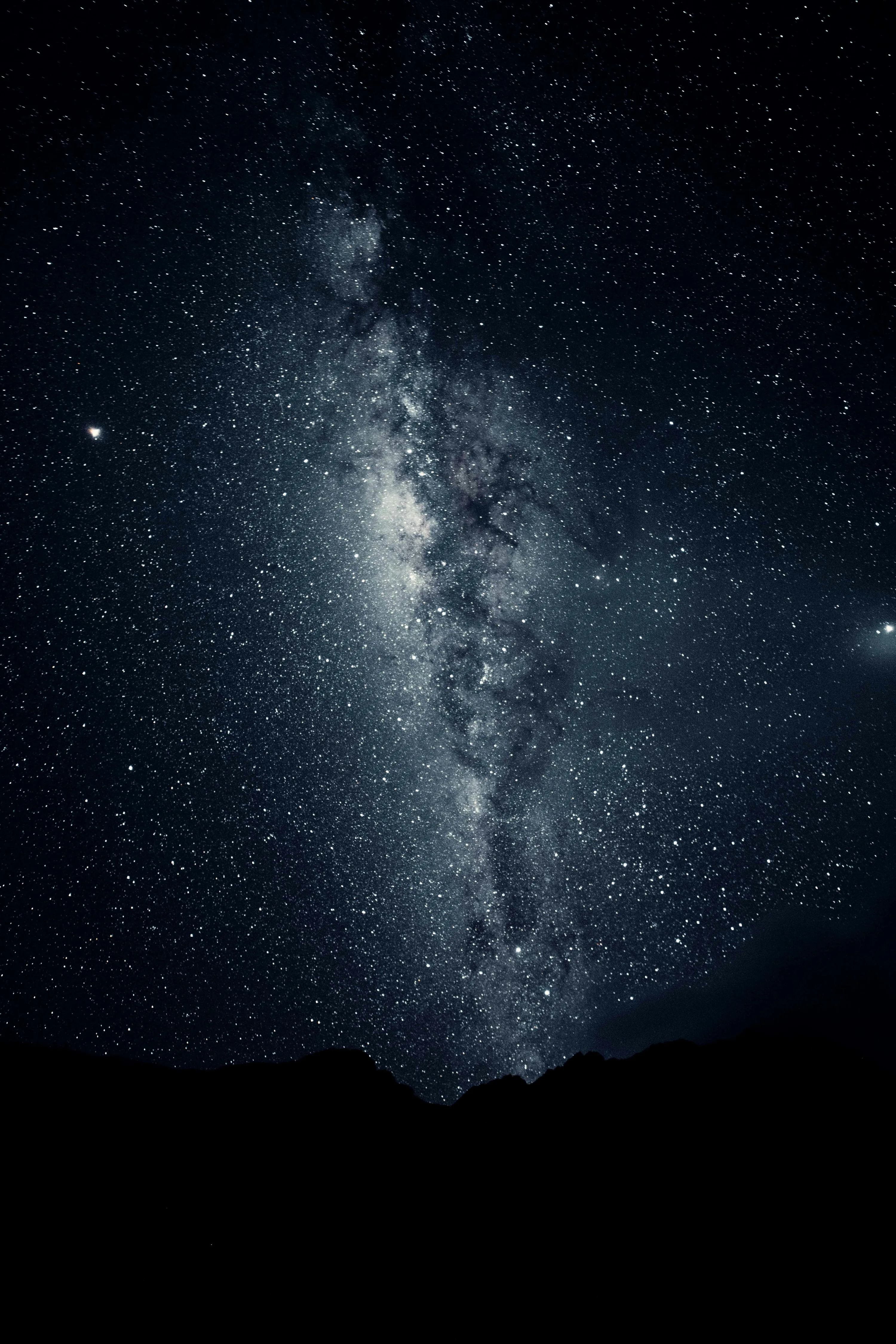 A vertical Milky Way column rises over a black mountain ridge in near-monochrome blue, a bright planet flaring on the left edge.