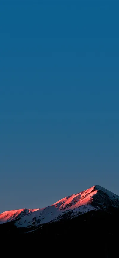 Late sun catches snow along a low ridge in red and pink while a clean blue gradient fills the tall sky above it.
