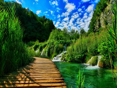 Wooden planks lead from the bottom corner past travertine falls spilling into clear green water, with a strip of clouded blue sky along the top.