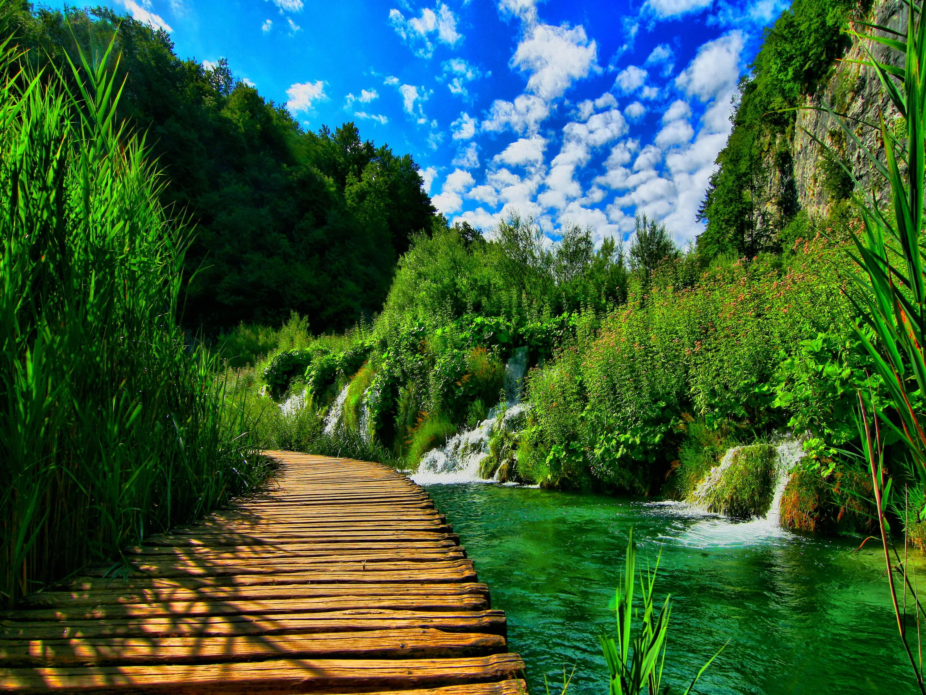 Wooden planks lead from the bottom corner past travertine falls spilling into clear green water, with a strip of clouded blue sky along the top.