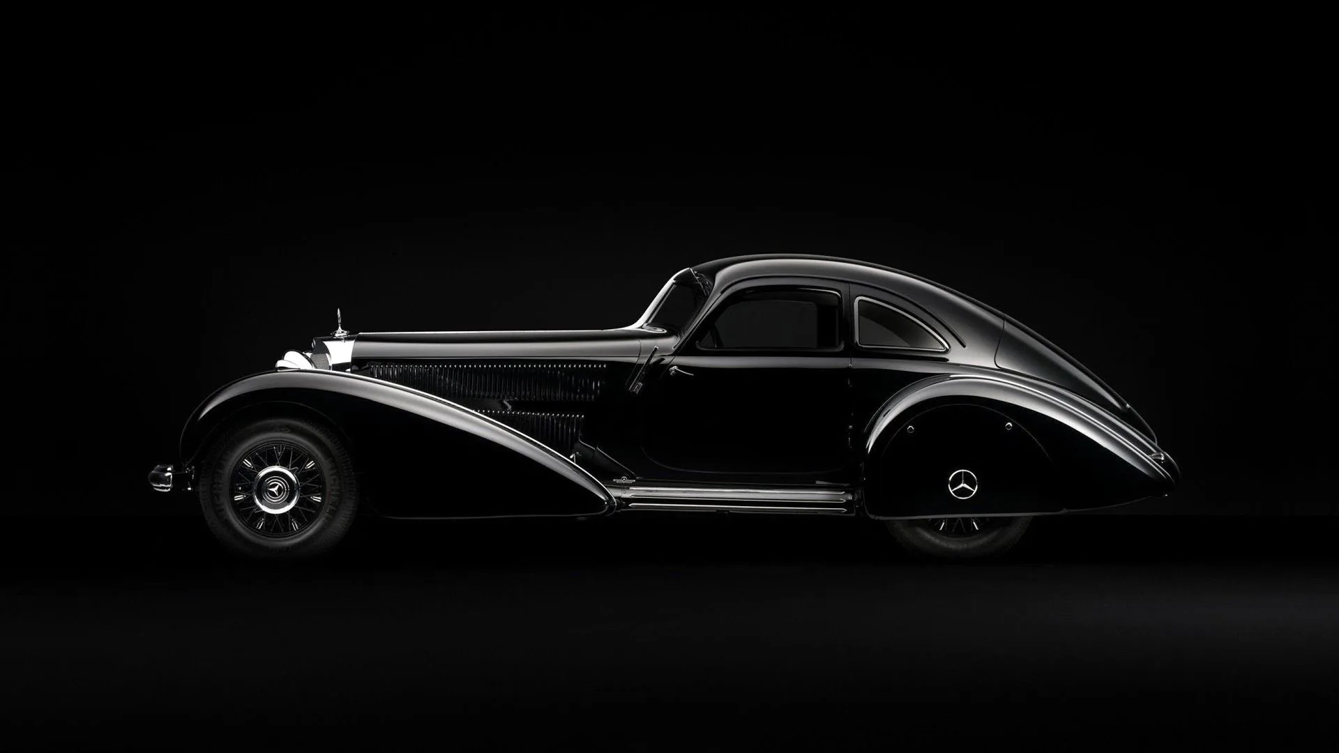 A pre-war aerodynamic coupe photographed side-on against pure black, chrome trim tracing its long teardrop roofline and covered rear wheel arch.