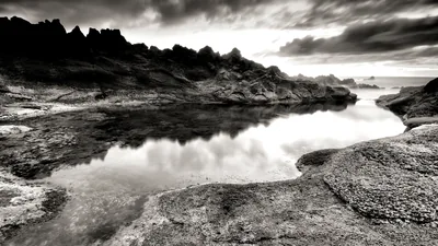 A still tidal pool mirrors the clouds between jagged coastal rocks, the whole scene pushed to hard monochrome with the sky brightest at the right edge.