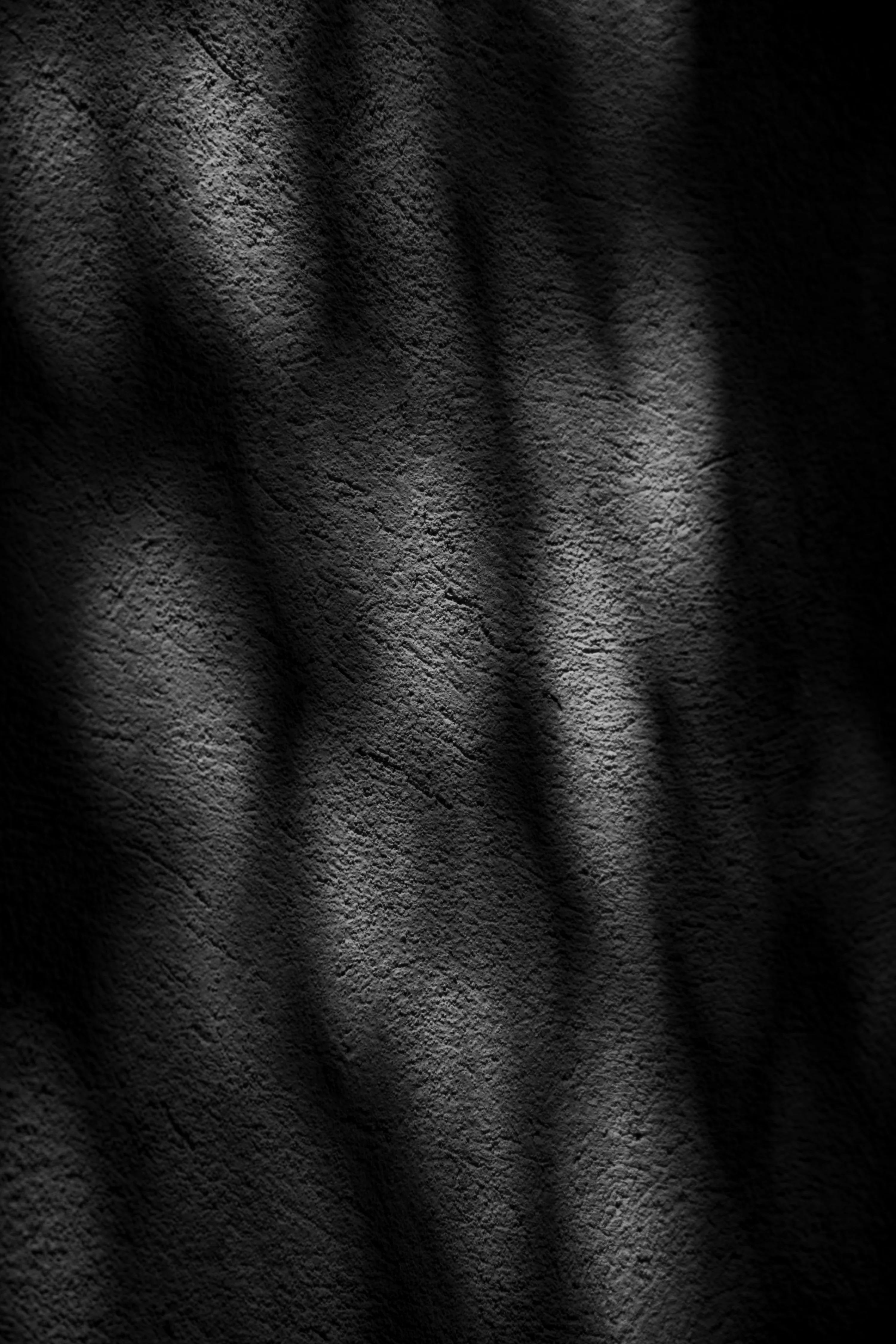 Soft vertical shadows fall across a coarse dark wall, the grain visible only where light grazes it.