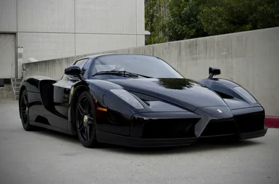 A gloss black Enzo parked on worn asphalt before pale concrete buildings, shot low from its front left, the bright wall keeping the frame's upper edge light.