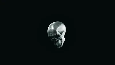 A small metallic skull hangs alone near the middle of an almost entirely black frame, its dented surface catching a weak grey light.
