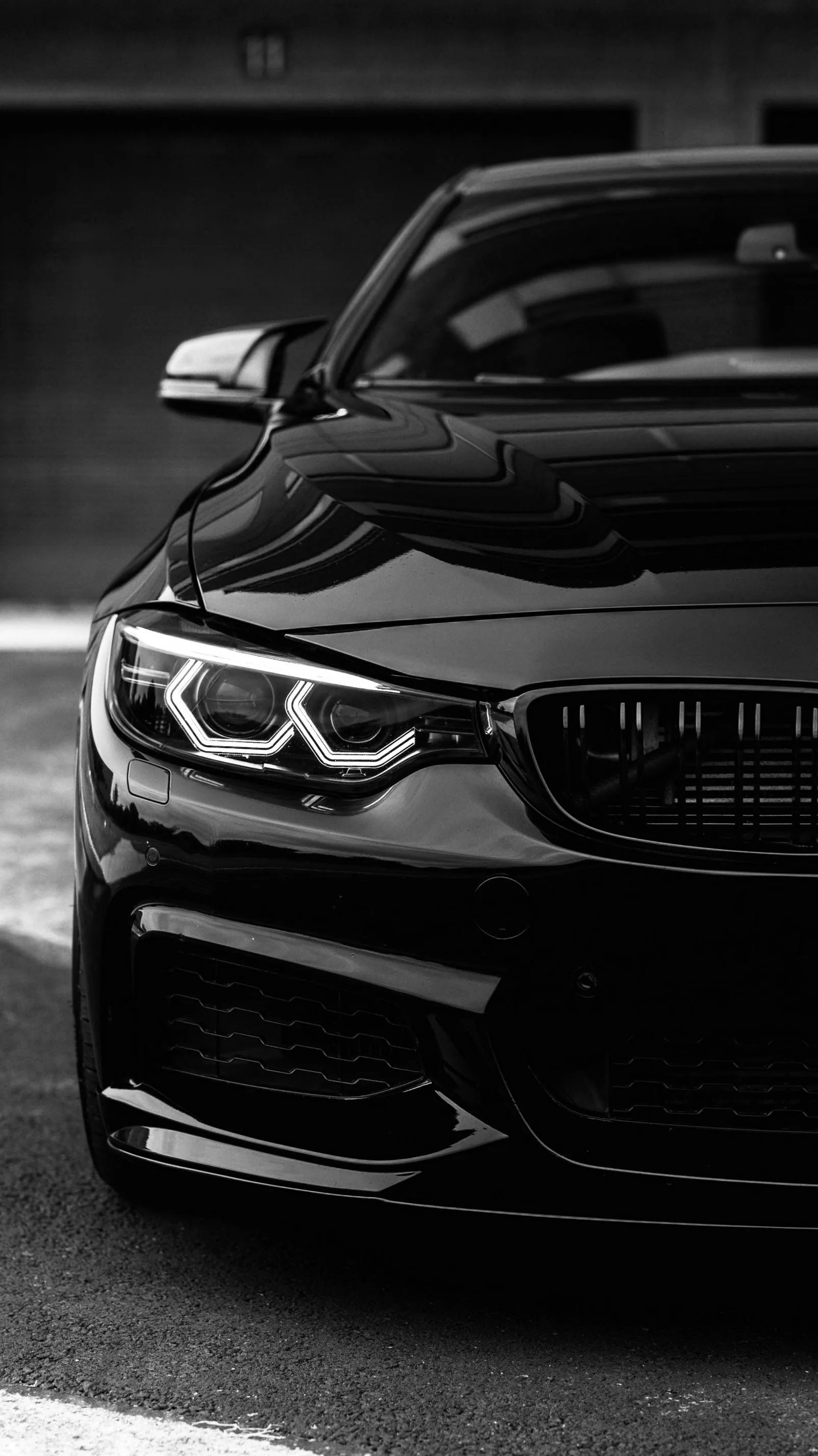A glossy black coupe photographed head-on in monochrome, the angel-eye headlight ring and kidney grille the brightest points in the frame.
