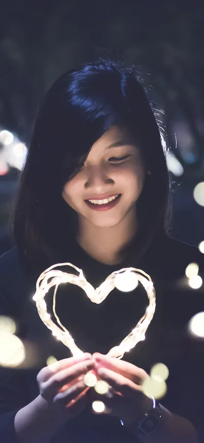 A smiling girl cups a heart bent from warm fairy lights, the night behind her breaking into orange bokeh.
