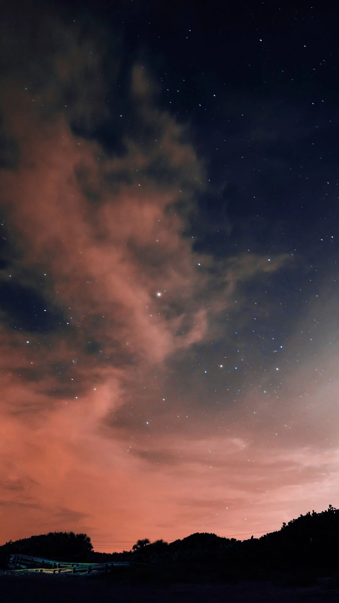 Reddish cloud drifts through a starfield above a black treeline, a pale glow rising along the right edge of the frame.