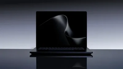 A slim black laptop sits open on a mirrored surface, a silky dark ribbon curling across its screen while the grey wall behind fades to black at the edges.