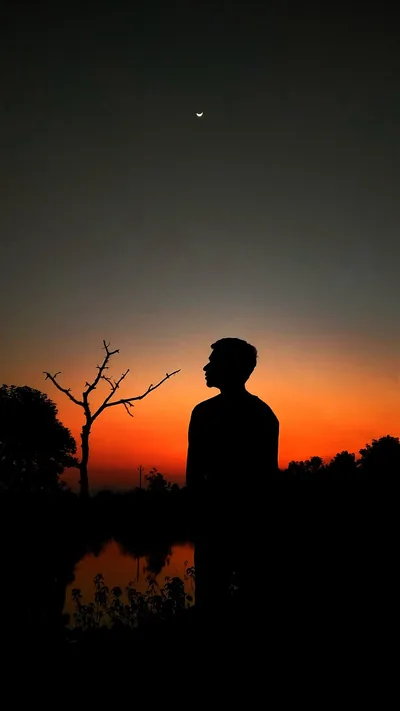 Deep orange burns along the horizon behind a young man gazing upward in profile, a bare tree at left and one faint point of light high in the black sky.