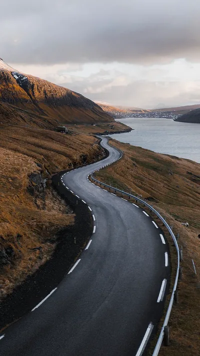 An empty two-lane road snakes down the right side toward distant houses across the water, low sun grazing the slopes from the left in this portrait shot.