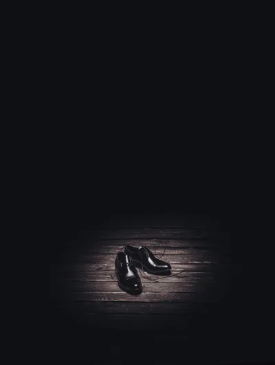 A pair of polished black leather shoes sits in a small pool of light on worn floorboards, the top half of the frame pure black for a lock-screen clock.