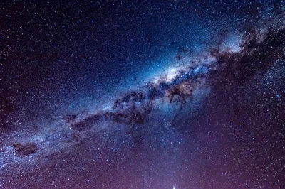 A long-exposure photograph of the galactic centre cutting diagonally across the sky, dust lanes splitting the blue-white glow.