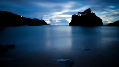 Long exposure turns the sea to blue mist around a tree-topped rock on the right, dark boulders surfacing across the smooth foreground water.