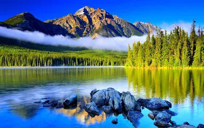 A red-brown pyramid of rock rises over saturated blue water, mist banding the treeline while smooth boulders break the surface in the foreground.