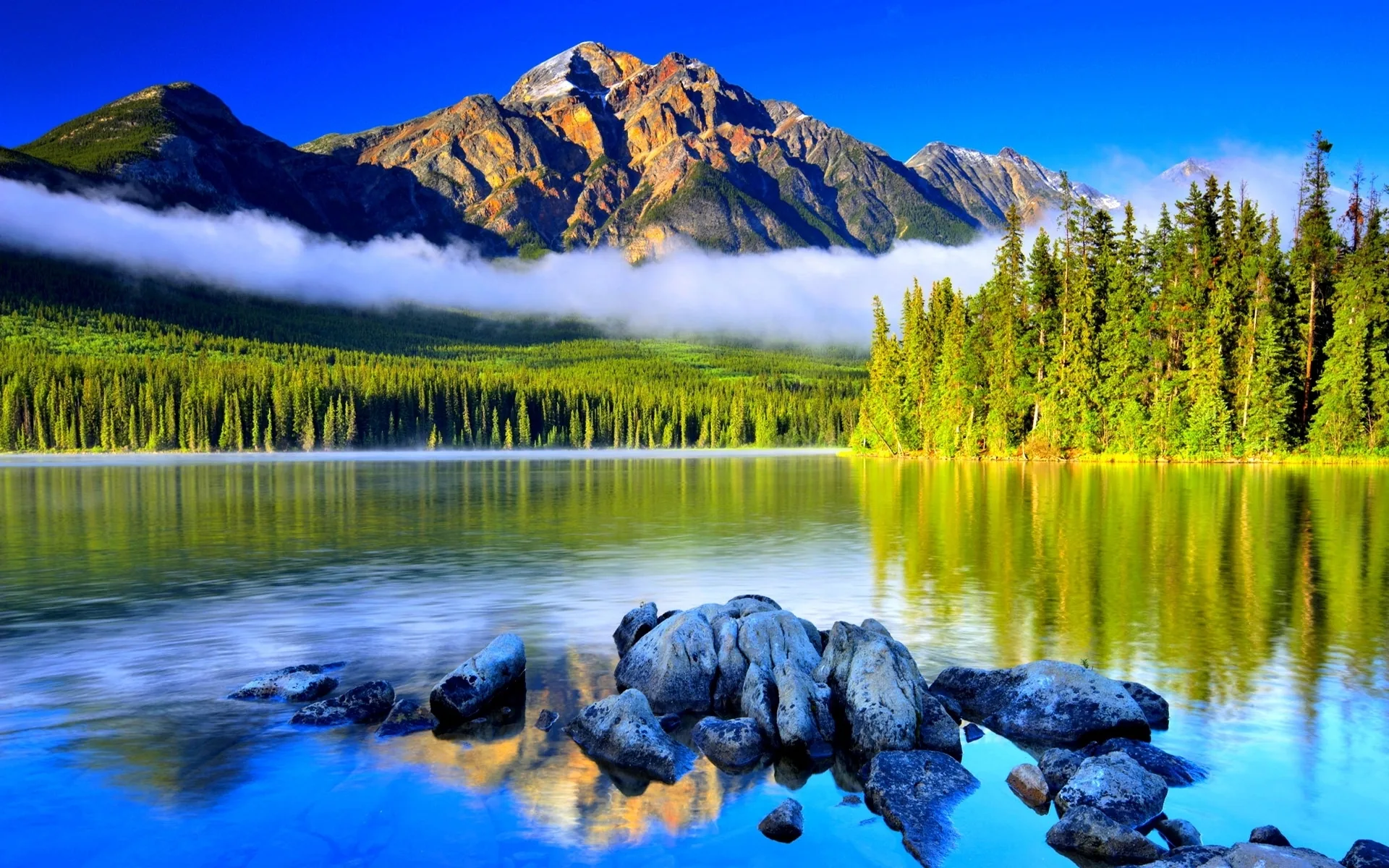 A red-brown pyramid of rock rises over saturated blue water, mist banding the treeline while smooth boulders break the surface in the foreground.