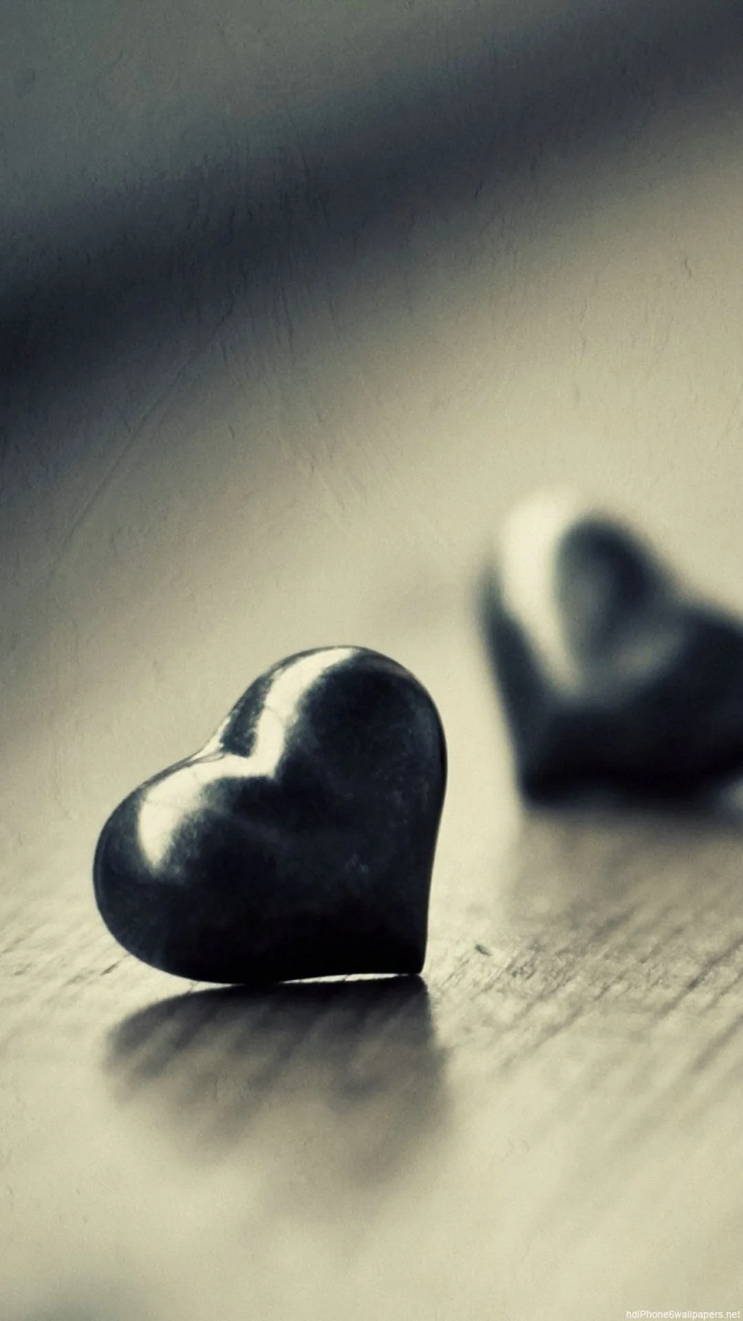 A polished black heart sits on a wooden surface in a tall crop, a second one out of focus behind it in muted grey-blue light.
