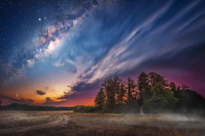 Streaked clouds sweep between the Milky Way and a sunset horizon, with mist drifting over a dirt track past dark trees below.