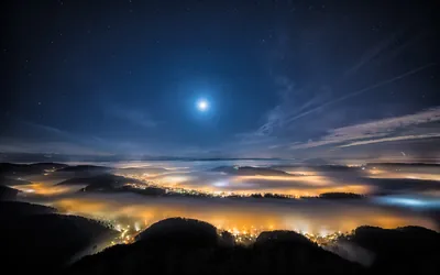 A bright moon hangs above a valley of fog at night, city lights burning gold and blue through the mist from below.