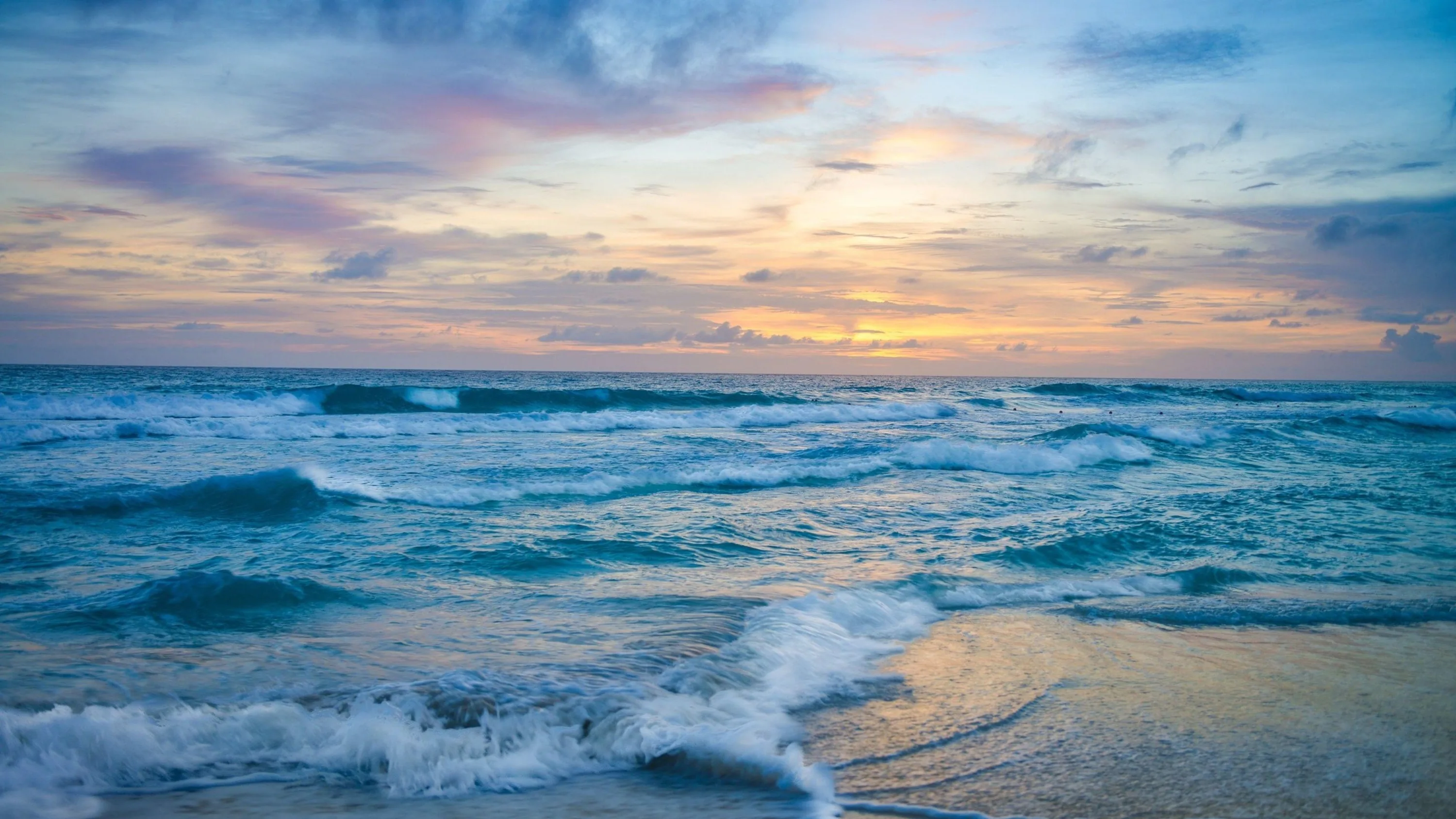 Small waves stumble ashore under pink and blue evening clouds, the soft-focus foreground and muted colours sitting quietly behind desktop icons.